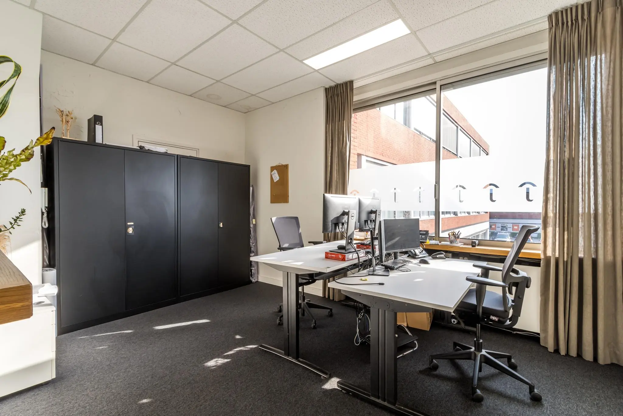 Modern kantoor met twee bureaus, bureaustoelen, computers en een groot raam met uitzicht op de Jan van Eycklaan.